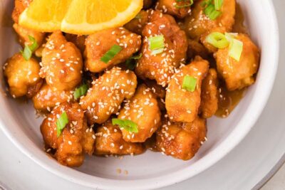 Keto orange chicken coated in a glossy orange sauce and topped with sesame seeds, green onions, and fresh orange slices.