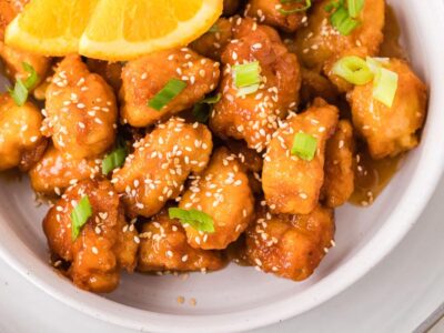 Keto orange chicken coated in a glossy orange sauce and topped with sesame seeds, green onions, and fresh orange slices.