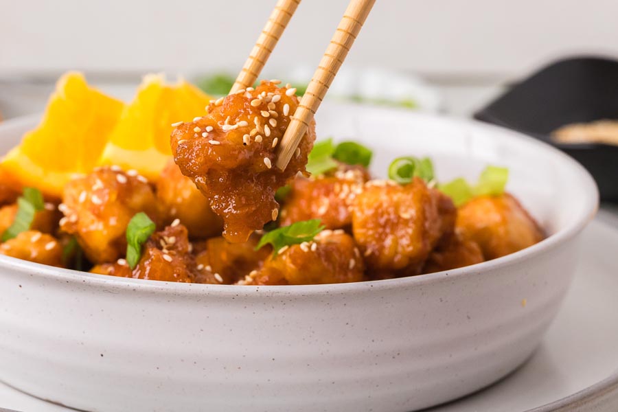 A piece of crispy keto orange chicken being lifted with chopsticks from a bowl garnished with sesame seeds, green onions, and orange slices.