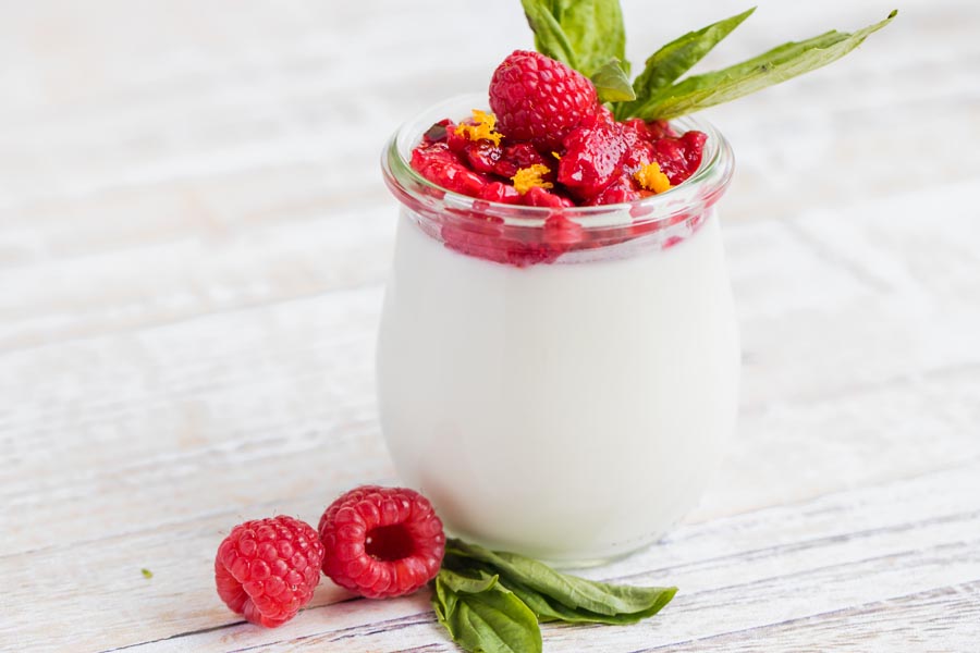 two sprigs of basil next to a creamy white panna cotta in a small jar with raspberries on top