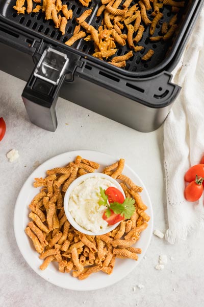 a bowl of baked pasta sits in front of an air fryer basket with more baked pasta chips inside