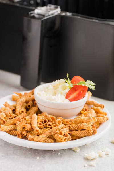 a plate of pasta chips with a dip in the center with an air fryer basket in the background