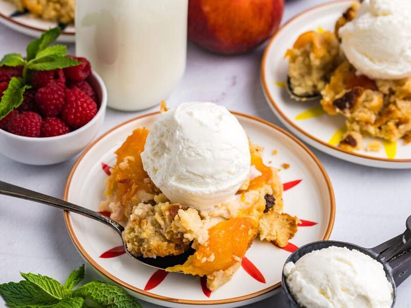 A spoon holding some peach cobbler on top of plate with fresh raspberries nearby.
