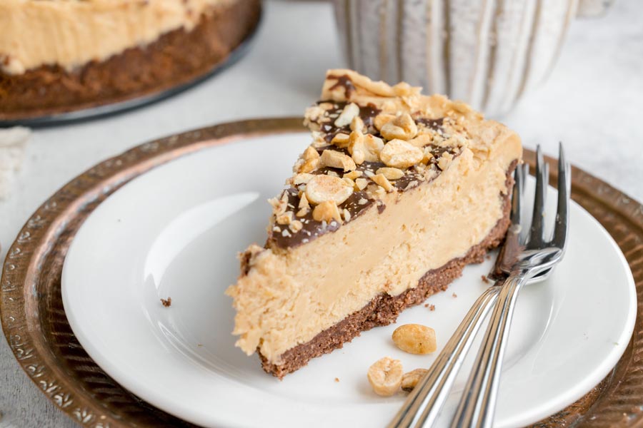 Slice of keto peanut butter cheesecake with chocolate crust, chocolate topping and chopped peanuts.