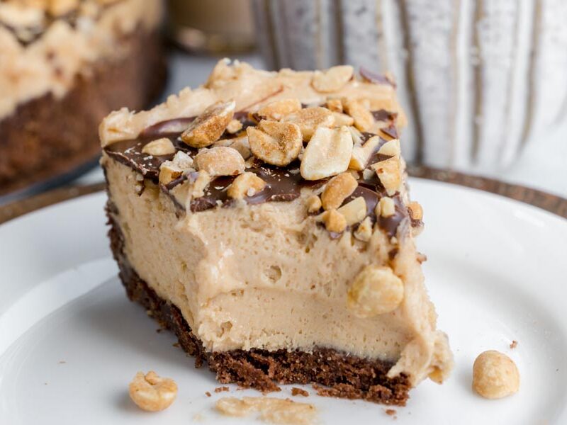 Thick slice of keto peanut butter cheesecake topped with chocolate and crushed peanuts on a dessert plate.