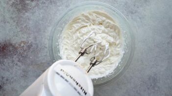 Whipped cream being mixed in a glass bowl with a hand mixer for the cheesecake filling.