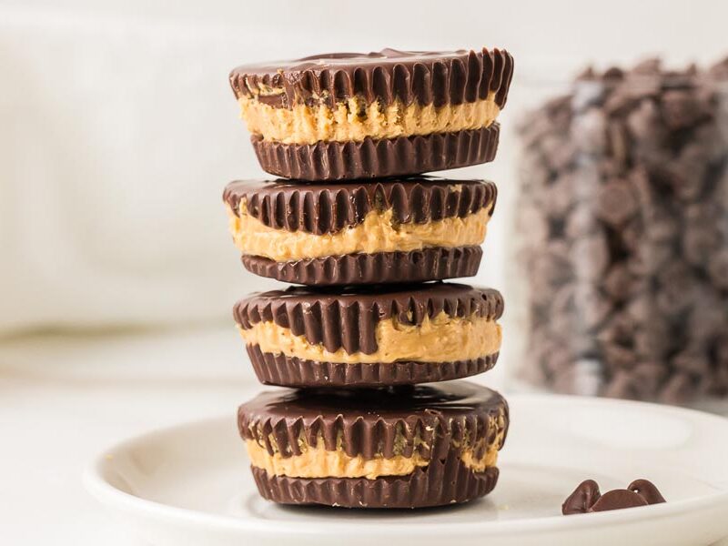 Four keto peanut butter cups stacked on a white plate with chocolate chips in the background.