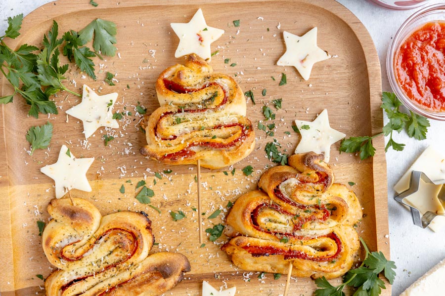 Three puff pastry Christmas trees on a wooden board next to pizza sauce, cheese stars and chopped parsley.