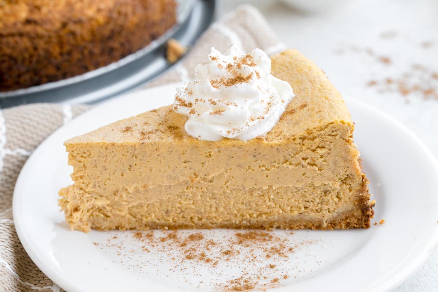 A slice of keto pumpkin cheesecake topped with whipped cream and sprinkled with cinnamon on a white plate.