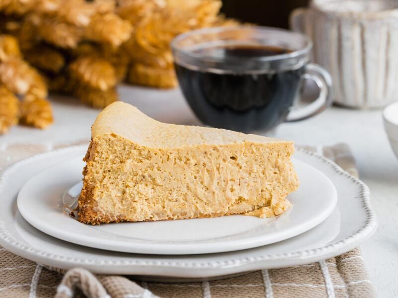 A plain slice of keto pumpkin cheesecake served on a white plate with a background of fall decor and a cup of coffee.