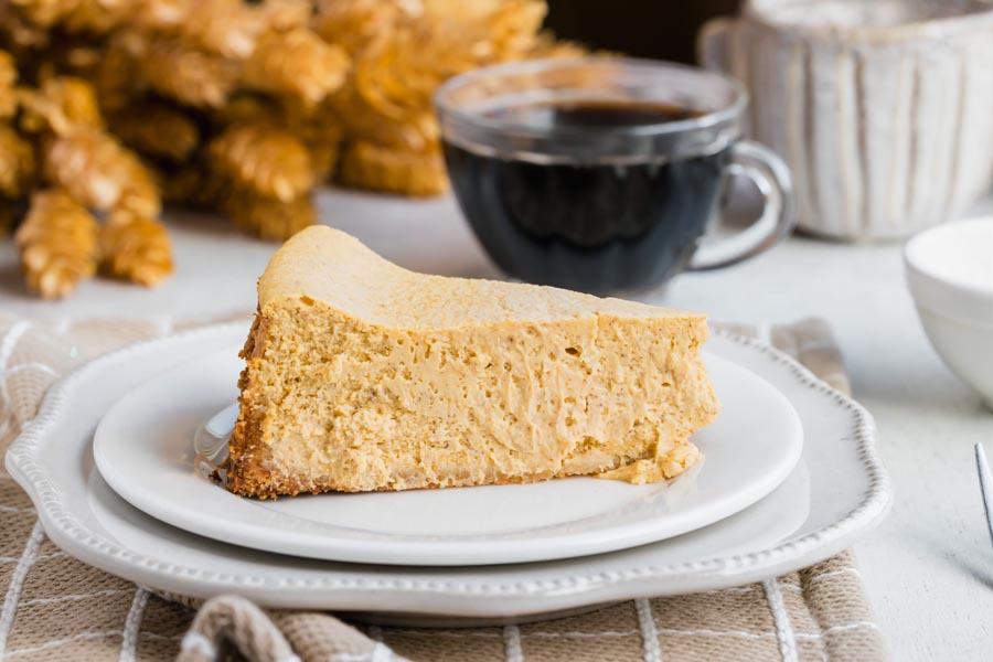 A plain slice of keto pumpkin cheesecake served on a white plate with a background of fall decor and a cup of coffee.