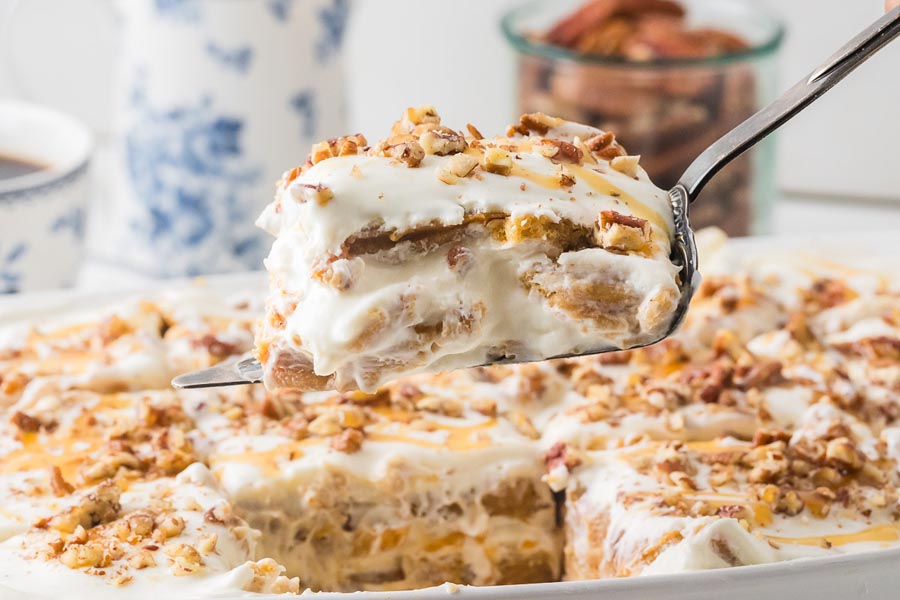 A spatula lifts a slice of pumpkin cheesecake lasagna from a dish, showing thick layers of pumpkin and creamy filling with pecan topping.