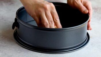 A hand pressing down into a greased springform pan to prepare it for the crust.