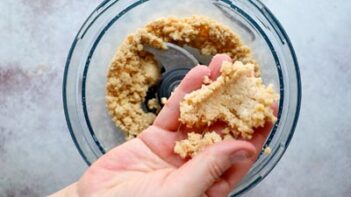A hand holding a clump of crumbly crust mixture above a food processor bowl.