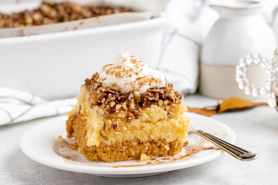 A slice of pumpkin cake sits on a white plate next to fork with whipped cream dusted with pumpkin pie on top.