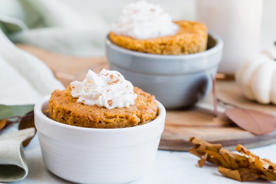 small pumpkin cakes coming out of ramekins topped with whipped cream and dashes of pumpkin pie spice with a white pumpkin in the background