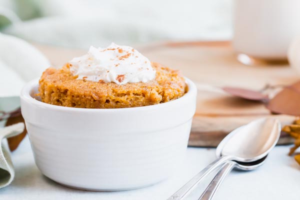 fluffy pumpkin flavored single serve cake in a white ramekin on a brown cutting board topped with a dollup of whipped cream