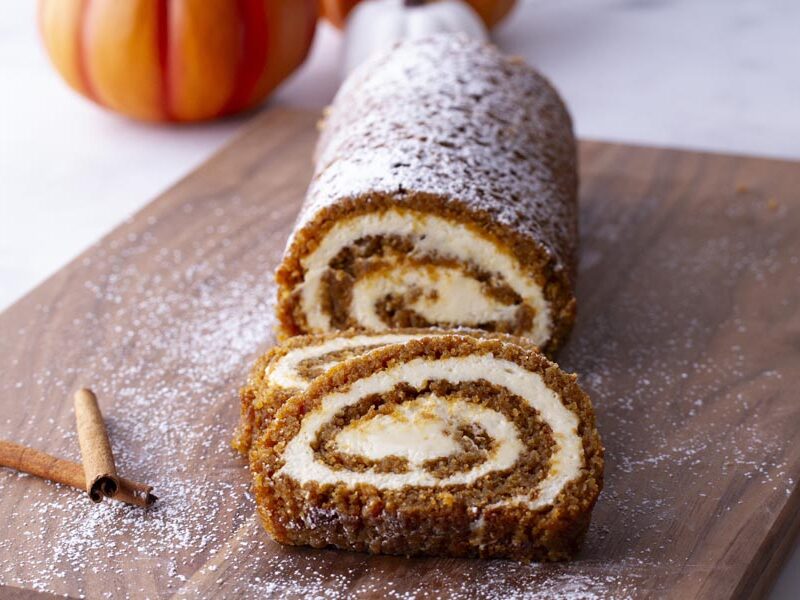 A pumpkin roll sliced on a cutting board with cinnamon sticks and powdered sweetener nearby.