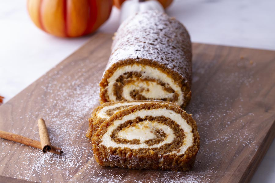 A pumpkin roll sliced on a cutting board with cinnamon sticks and powdered sweetener nearby.