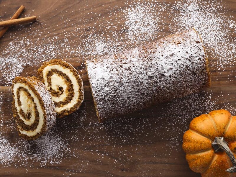 A pumpkin roll cake with no cracks on a cutting board with a pumpkin nearby and two slices of cake.