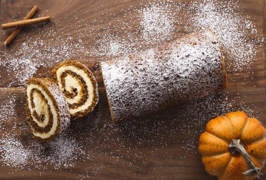 A pumpkin roll cake with no cracks on a cutting board with a pumpkin nearby and two slices of cake.