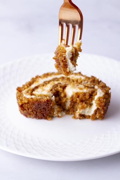 A fork holding a bite of pumpkin cake over a plate with a slice of pumpkin roll on it.