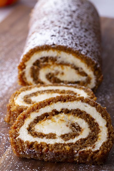 A pumpkin cake rolled up with cream cheese filling to form a pumpkin roll. Two slices sit in front of the cake roll.