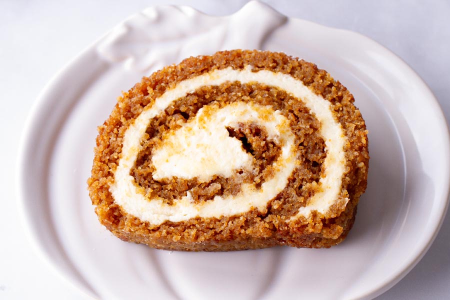 A white pumpkin plate with a slice of pumpkin roll spiraled with cream cheese filling on it.