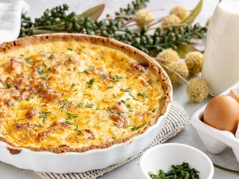 a quiche in a white tart dish with a crust next to milk, eggs and a bowl of chives
