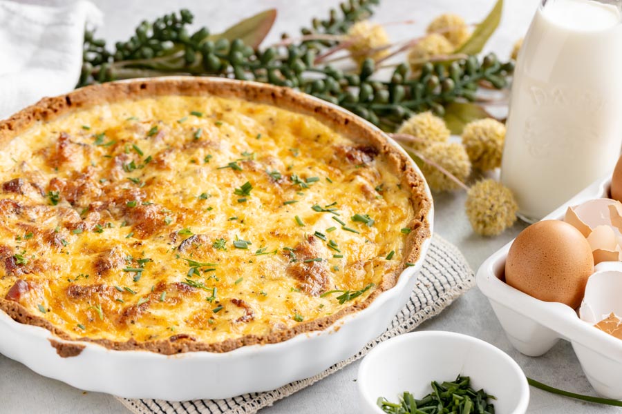 a quiche in a white tart dish with a crust next to milk, eggs and a bowl of chives