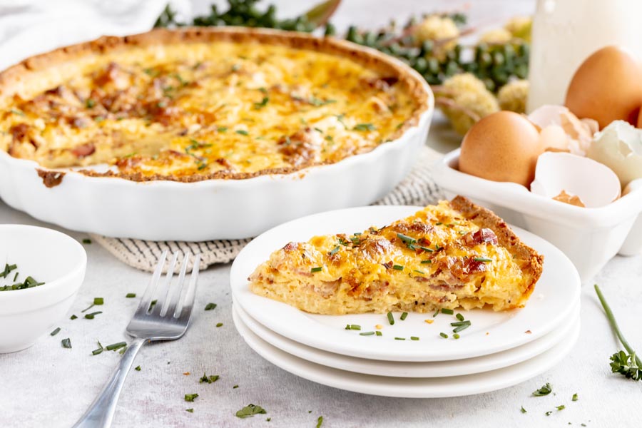 a slice of quiche on a stack of plates next to a fork with parsley sprinkled around