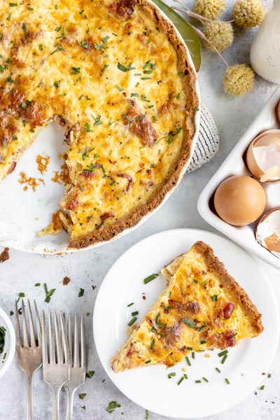 a slice of quiche lorraine on a dish with the whole quiche above and forks nearby