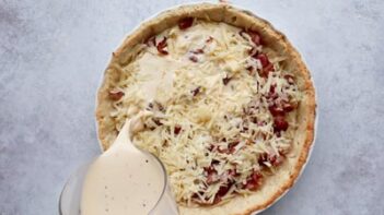 pouring egg mixture into a pie crust filled with shredded cheese and bacon