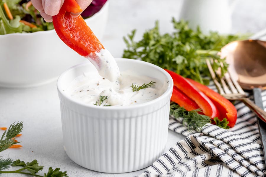 dipping a red bell pepper slice into a bowl of ranch dressing with more sliced pepper behind and a spoon and fork as well