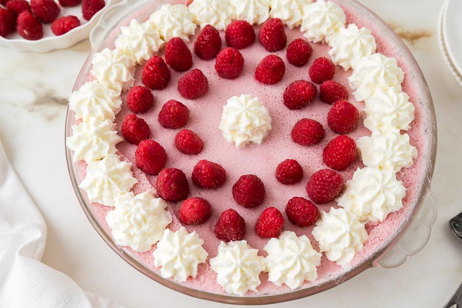 A top-down view of a no-bake keto raspberry marshmallow cream pie garnished with piped whipped cream and fresh raspberries.
