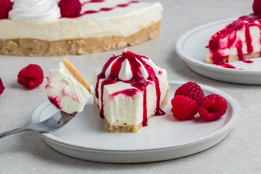 A bite of cheesecake on a fork in front of a slice of raspberry cheesecake on a plate.