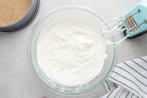 A glass bowl with cheesecake mixture inside next to an electric mixer.