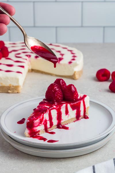 Pouring raspberry sauce over a slice of cheesecake topped with fresh raspberries.