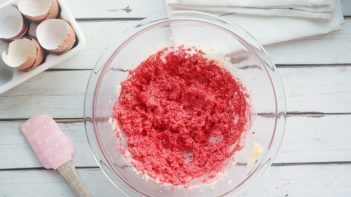 broken egg shells sit by a red batter in a bowl