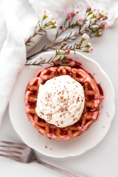 a stack of red waffles topped with whipped cream next to a fork and pink flowers