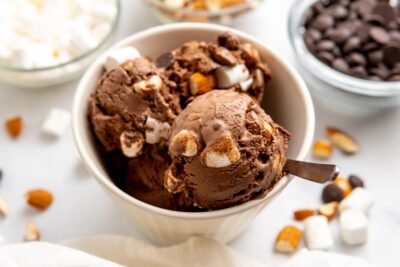 A bowl of keto rocky road ice cream with a chocolate base and chunks of almonds, chocolate and mini marshmallows inside. Toppings like nuts and chocolate are scattered around the bowl.