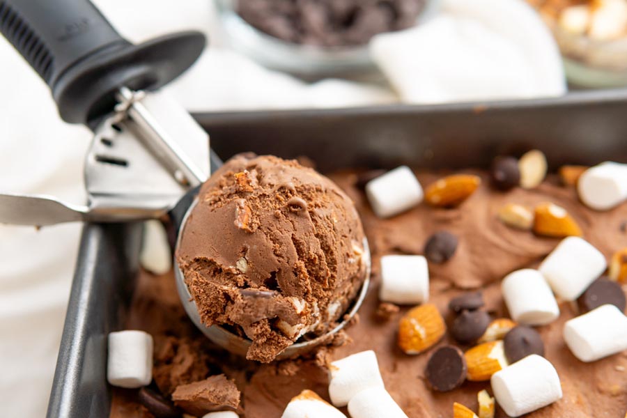 A scoop holding a ball of frosty, creamy chocolate ice cream with chunks of nuts and marshmallows. Whole nuts, chocolate chips and marshmallows sit on top of the tub of swirly ice cream.