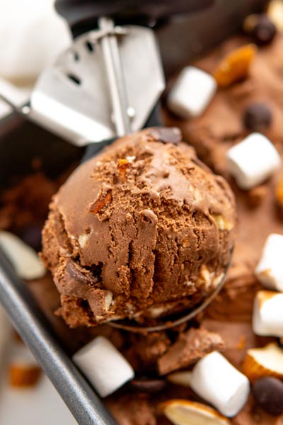 An ice cream scoop with a ball of rocky road ice cream in it. Mini marshmallows and nuts lie on top of the ice cream container below.