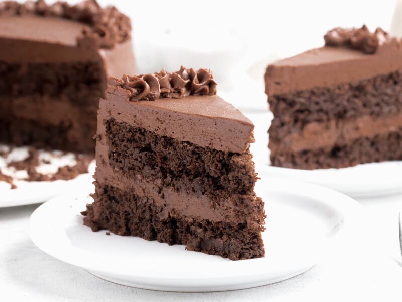 A slice of chocolate cake with chocolate frosting on a plate with another slice behind.