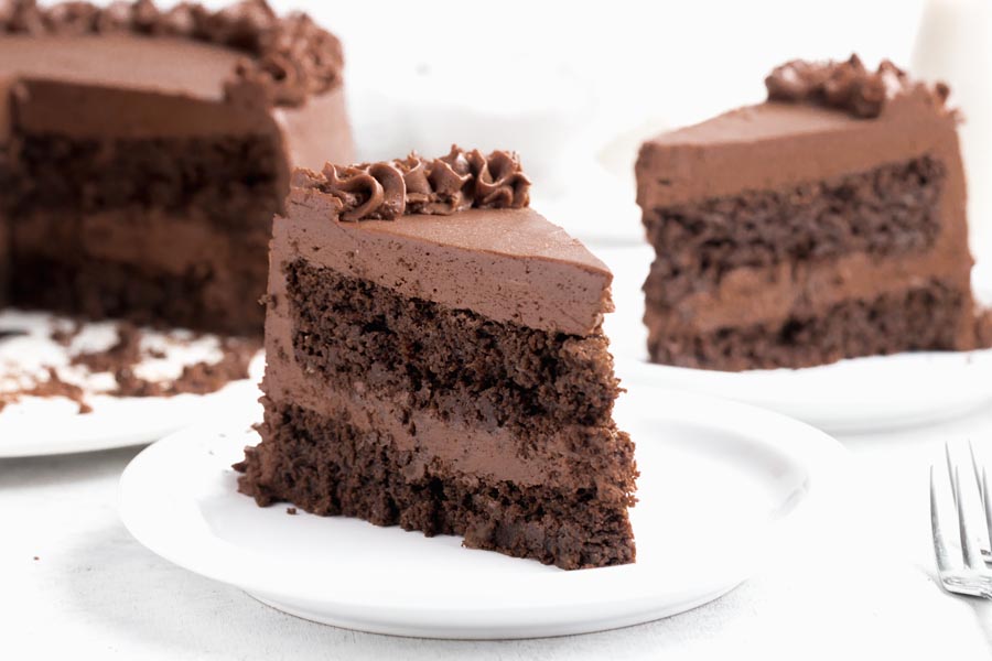 A slice of chocolate cake with chocolate frosting on a plate with another slice behind.