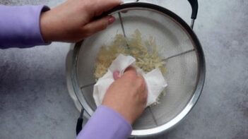 Hands pressing water out of shredded sauerkraut through a strainer with a paper towel.