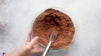 A hand holding a bowl with dry ingredients containing cocoa with a whisk inside.