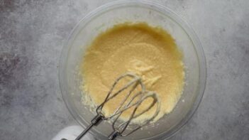 Electric mixer beaters are above a clear bowl with butter and eggs mixed inside.