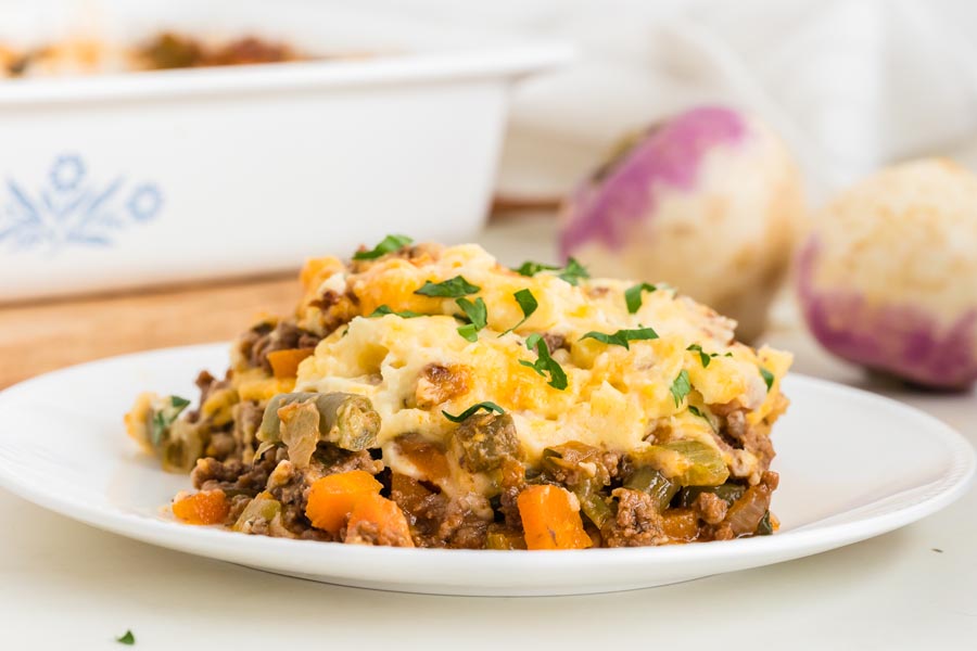 A serving of shepherd's pie on a white plate filled with greens and carrots with turnips in the background.