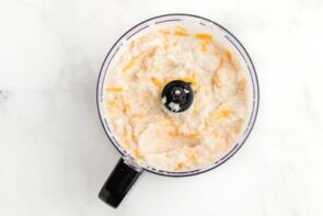 A food processor bowl with mashed turnips and shredded cheese inside.